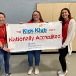 Three staff hold a Kids Klub banner reading Nationally Accredited, promoting EnrichEd programs and community support.