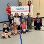 EnrichEd staff and children in a gym hold a Kids Klub Nationally Accredited banner, sharing community enrichment.