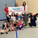 Kids and staff in a gym holding a Kids Klub Nationally Accredited banner, playful poses — EnrichEd program.