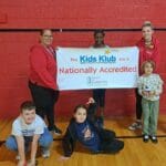 EnrichEd Kids Klub staff and children smiling while holding a 'Nationally Accredited' banner in a gym.