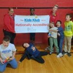 Children and staff hold a Kids Klub Nationally Accredited banner in a gym, showcasing EnrichEd program and safe community