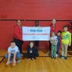 Kids and staff in a gym holding a 'Kids Klub Nationally Accredited' banner, EnrichEd after-school safety and enrichment.