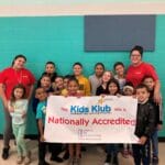 Smiling students and two staff hold a Kids Klub 'Nationally Accredited' banner in a bright enrichment space - EnrichEd.