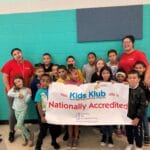 Children and staff holding a Kids Klub Nationally Accredited banner for EnrichEd's program in a safe community site.