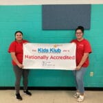Two staff in red shirts hold EnrichEd Kids Klub banner announcing the site's National Accreditation for enrichment programs.