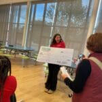 Smiling staff holding a Kids Klub award check in a school cafeteria, celebrating EnrichEd enrichment and community support