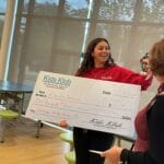 Smiling staff holding oversized Kids Klub check and receiving a trophy in a bright community room, EnrichEd.