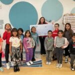 Smiling elementary students and staff pose with a large check in a bright EnrichEd program classroom, community support.