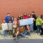 Smiling children and mentors holding a giant check and trophy by a brick wall, EnrichEd after-school support program