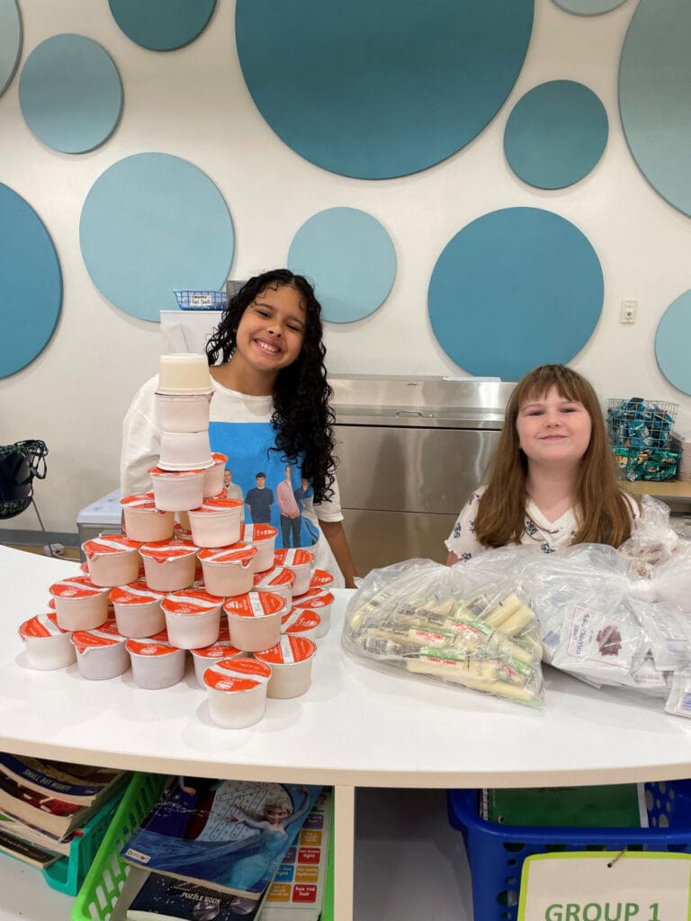 Child And Adult Food Care Program Announced 1 Two smiling students at an EnrichEd snack table stacking yogurt cups and packaged snacks under blue circular wall decor.
