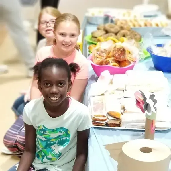 Three smiling children at an EnrichEd snack and craft table with sandwiches and pastries in a community enrichment setting.