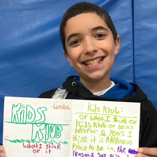 Smiling student holding handwritten Kids Klub poster highlighting EnrichEd after-school enrichment and community support.