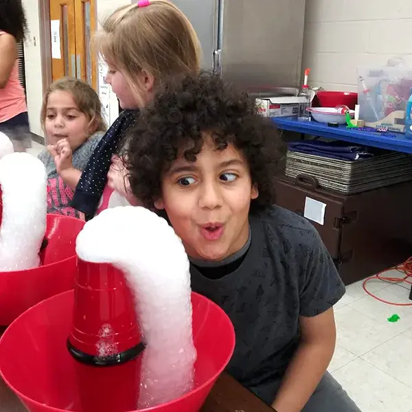 Children in EnrichEd program watch foam erupt from red cups during a hands-on science activity, boy reacts with delight.