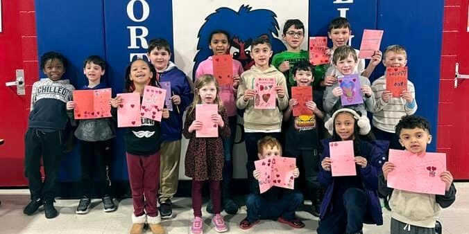 Elementary students holding handmade Valentine's cards in a school hallway, smiling together — EnrichEd community support.
