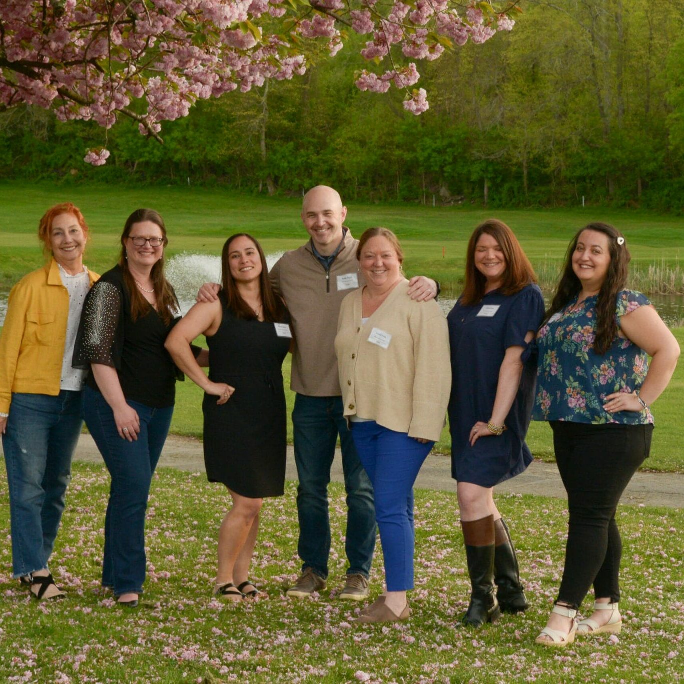 Seven people smiling under a blooming tree by a pond, EnrichEd team at an outdoor community enrichment event