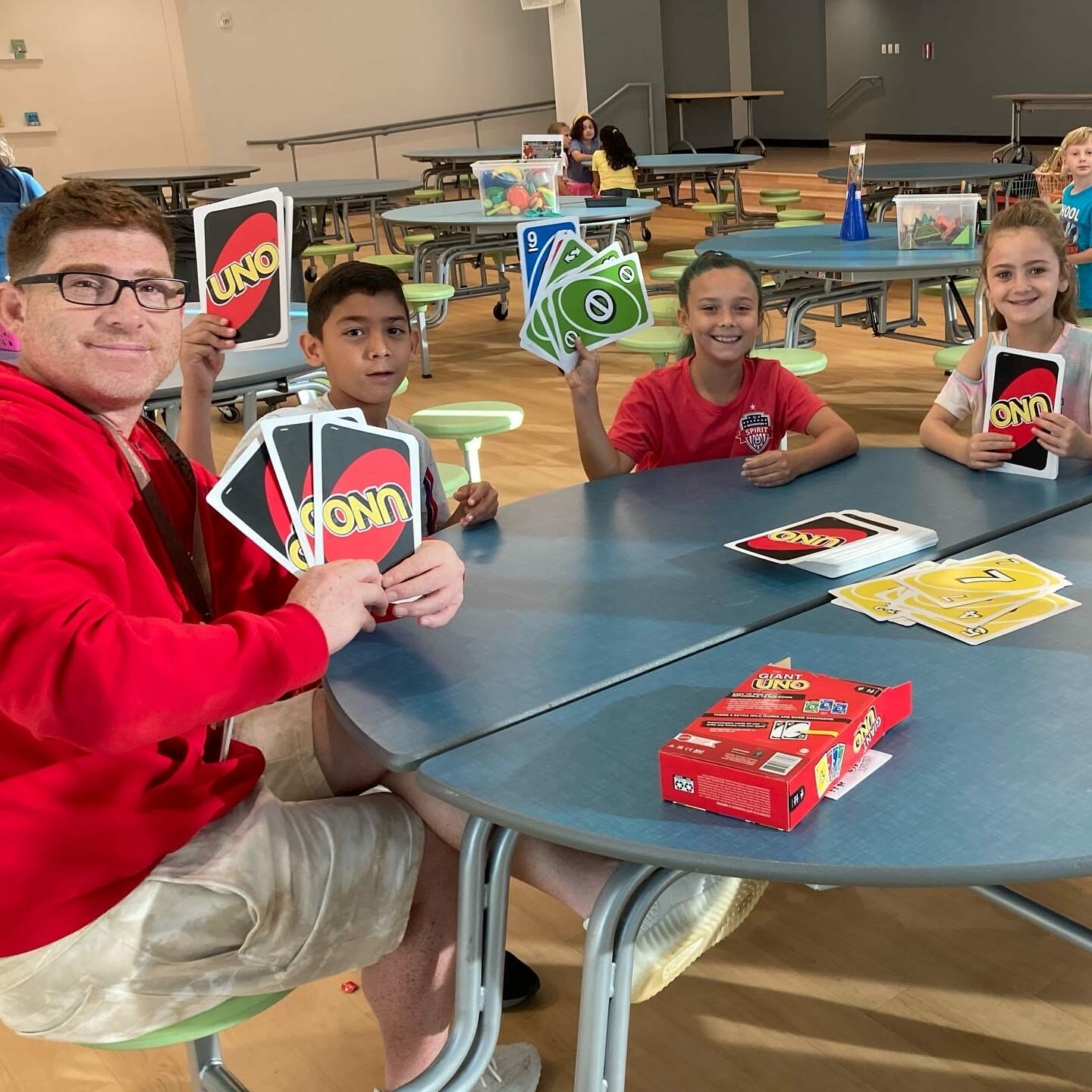 Adult and children play giant UNO at a cafeteria table, EnrichEd enrichment offering safe, supportive community activity
