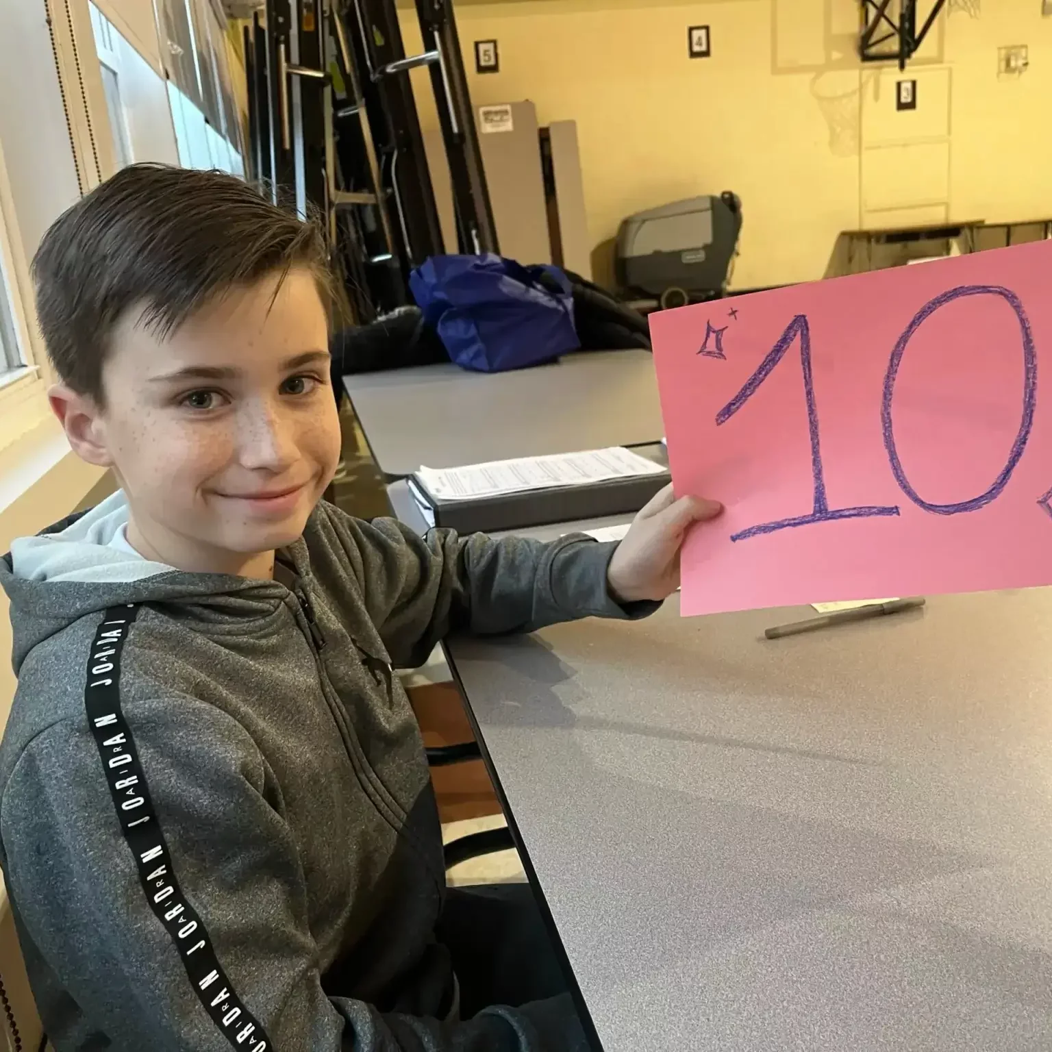Smiling student at EnrichEd holding pink sign reading '10' while seated at a gym table in a supportive enrichment setting.