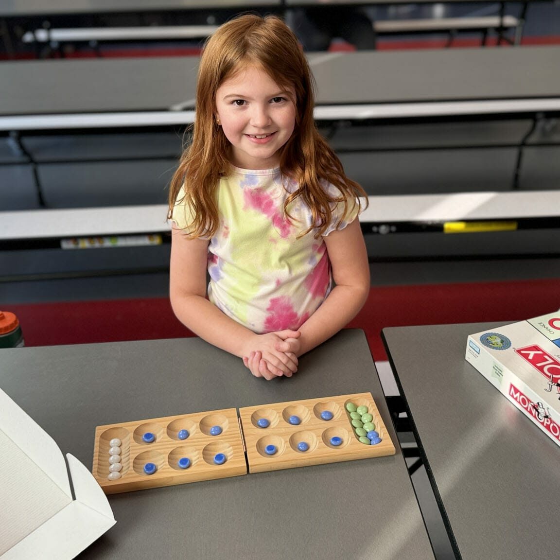 Smiling girl at a table playing mancala with Monopoly nearby, part of EnrichEd enrichment activities in a safe community hub
