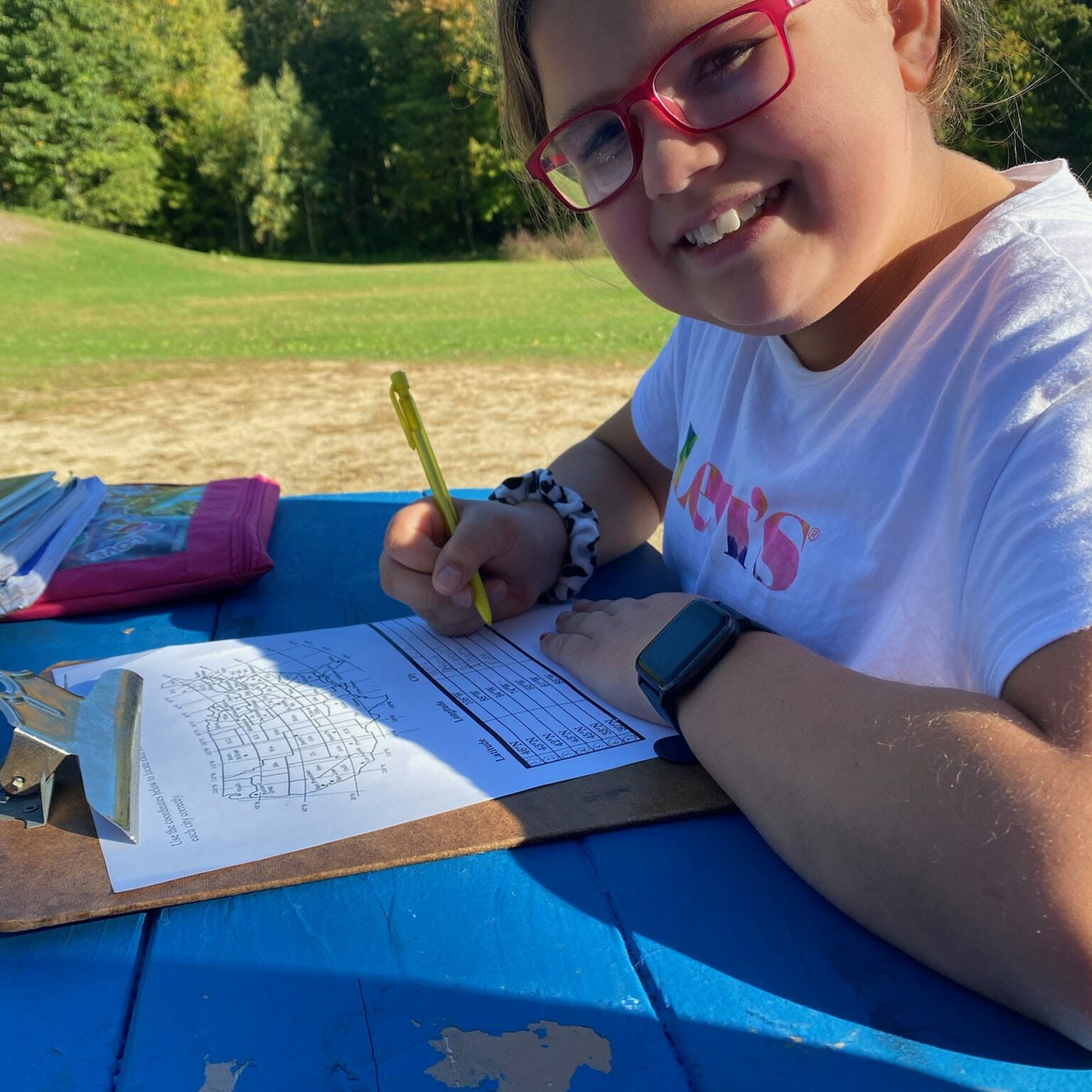 Smiling student with pink glasses writes on a worksheet at a blue picnic table outdoors, EnrichEd community program.