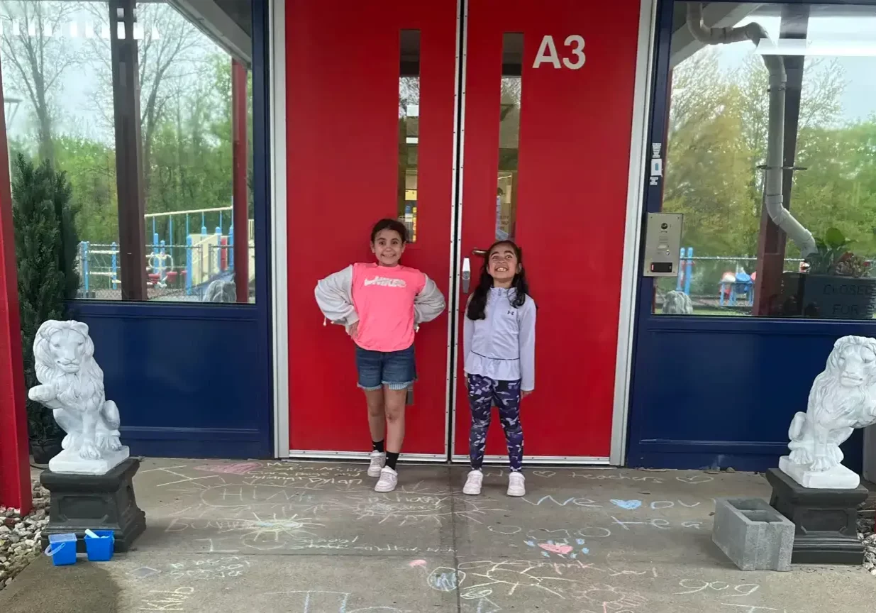Two smiling girls stand by red A3 doors with chalk art on the pavement and lion statues, EnrichEd after-school setting.