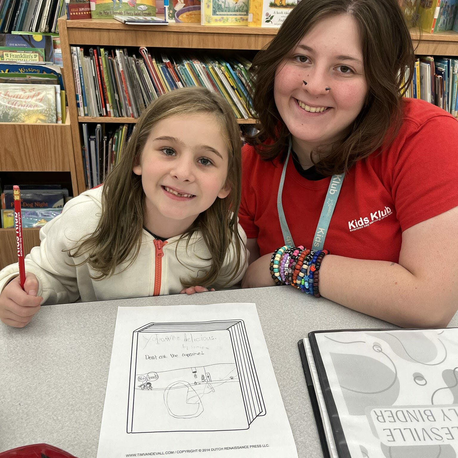 Smiling child and EnrichEd mentor at a library table with books, working on a drawing worksheet in a safe learning space.