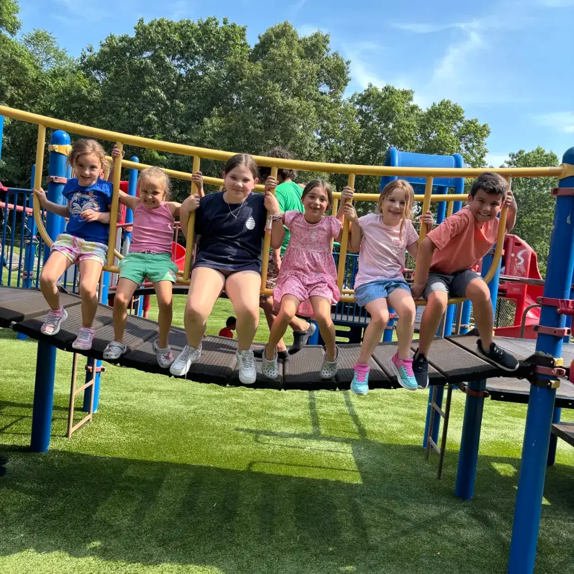 Seven children sit on a playground bridge holding a yellow rail, smiling under blue sky - EnrichEd community enrichment.