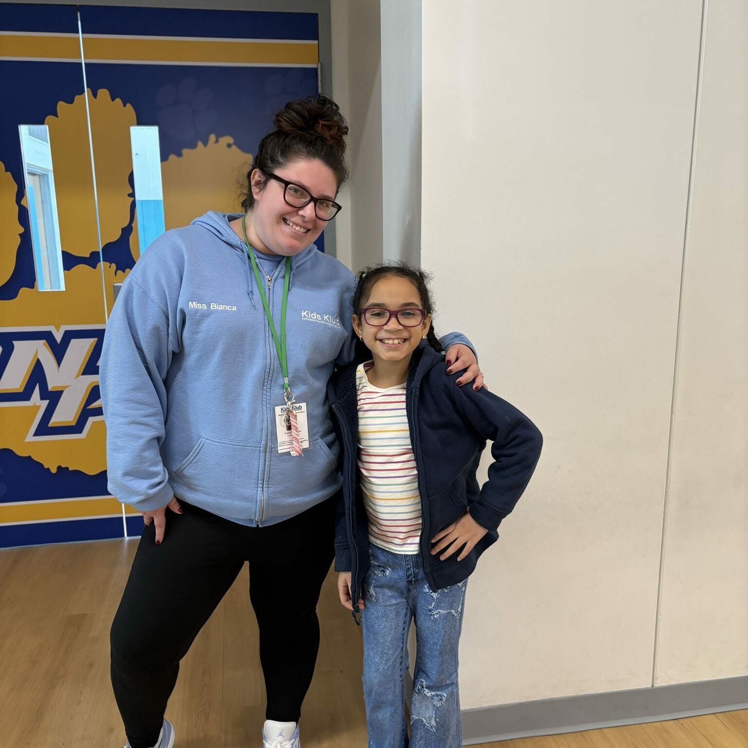 Smiling after-school staff member with arm around a smiling student in school hallway, EnrichEd after-school program.