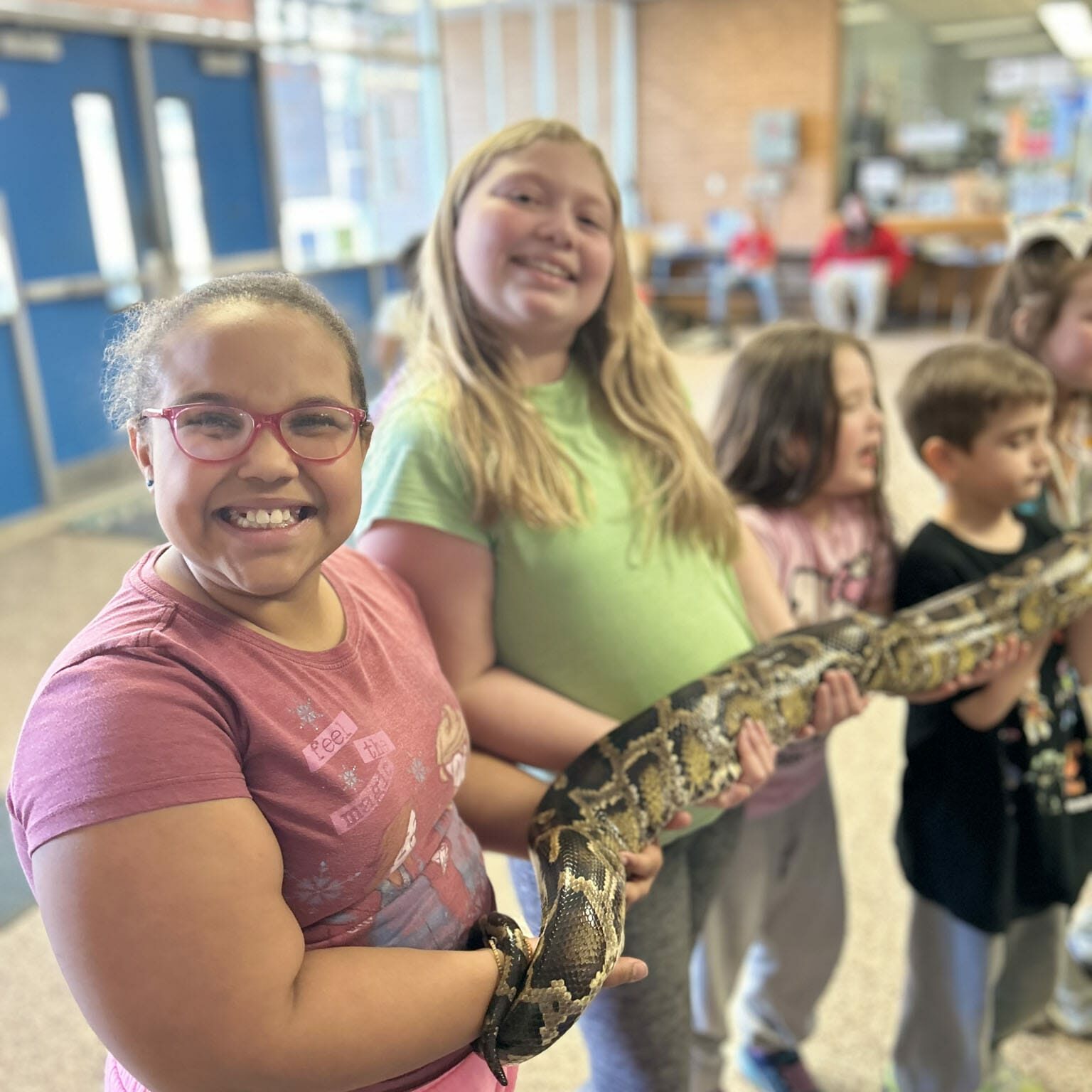 Smiling children holding a large snake in a school hallway during an EnrichEd enrichment activity, community learning.