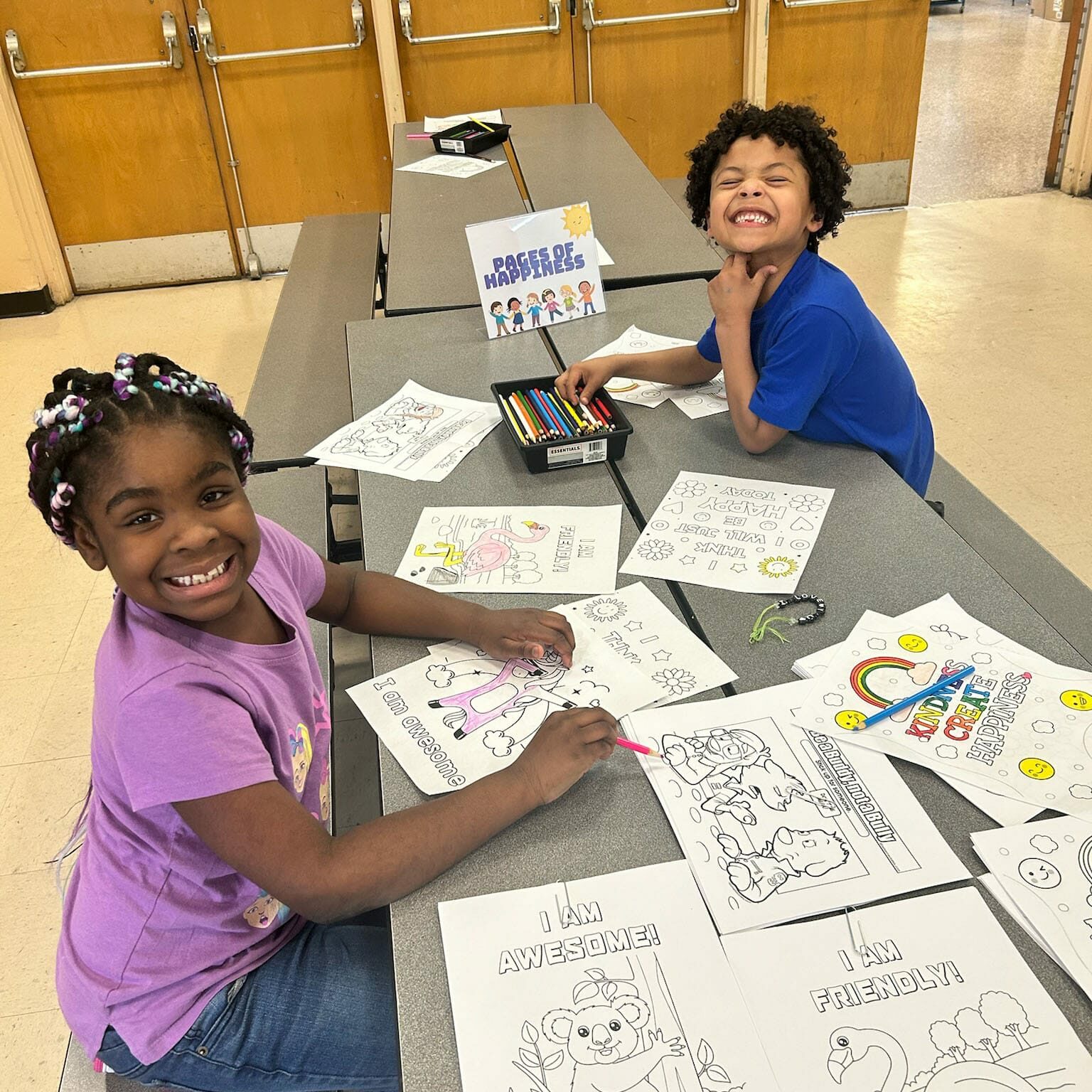 Two kids smiling and coloring positive worksheets at an EnrichEd enrichment table, promoting a safe, supportive space.