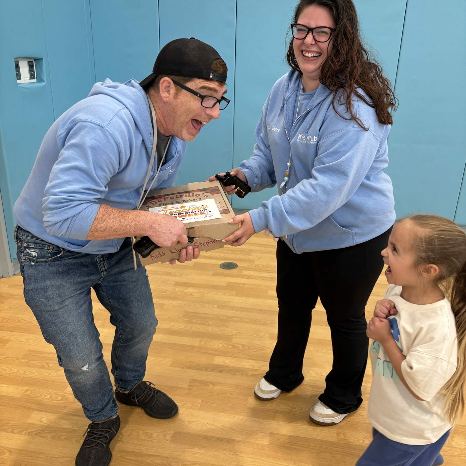 Two adults in light-blue hoodies laugh while handing a pizza box to a delighted child in a gym, EnrichEd program.