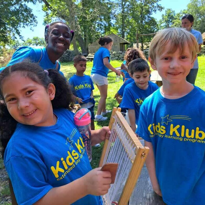 Children in blue Kids Klub shirts smiling outdoors, using a wooden loom in an EnrichEd enrichment activity