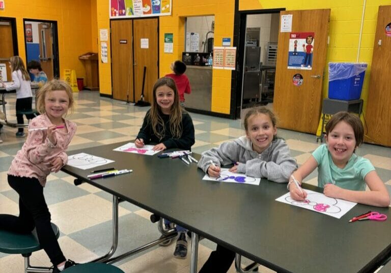 Four smiling children coloring at a cafeteria table with markers, yellow walls — EnrichEd afterschool program