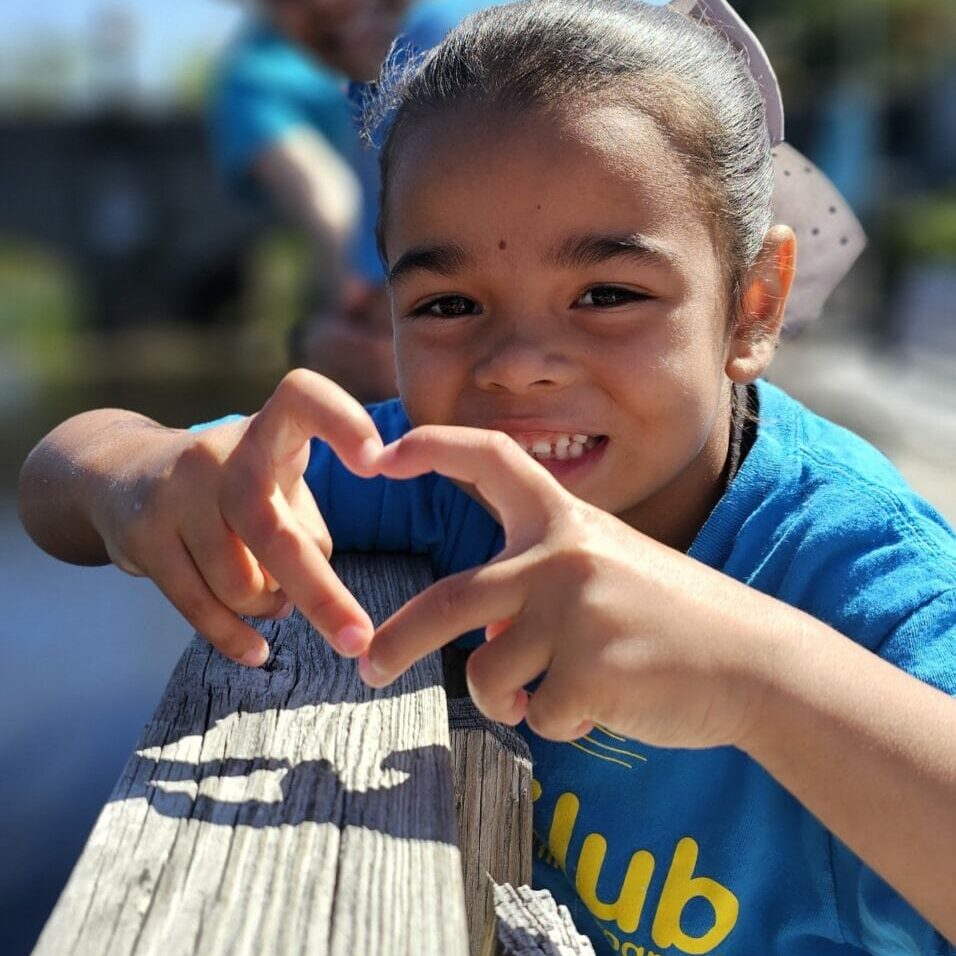 Smiling child making a heart with hands on a wooden railing, wearing a blue EnrichEd program shirt, peer blurred behind.