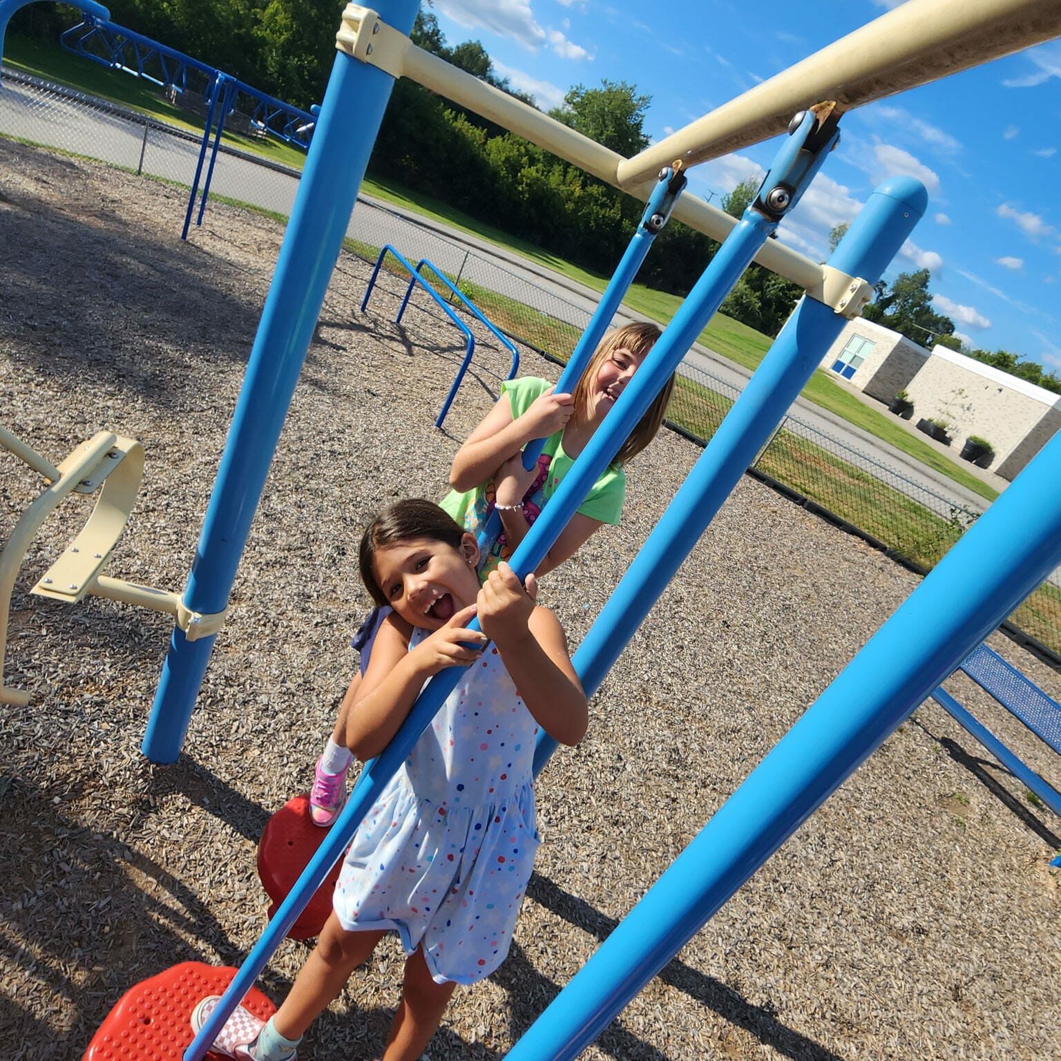 Two children smiling on blue playground poles by a school building, representing EnrichEd enrichment and community support.