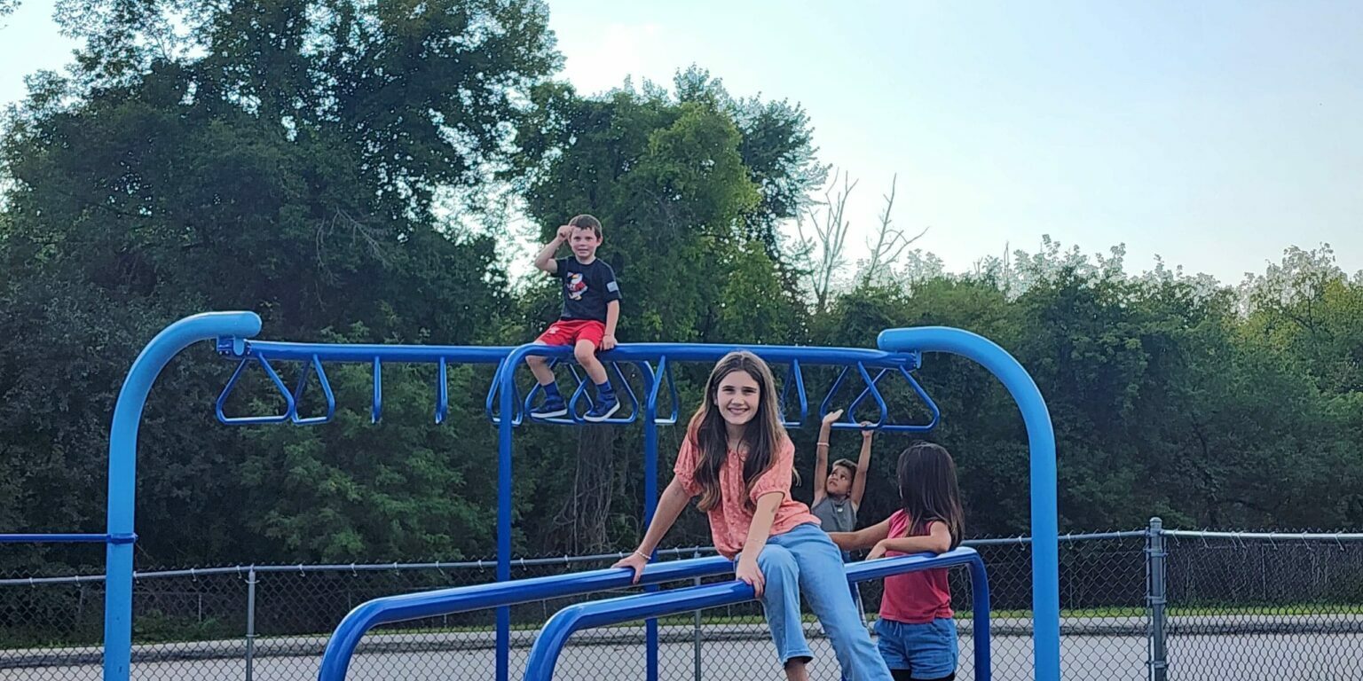 Children play on blue monkey bars at a park, smiling and climbing; EnrichEd supports safe, enriching community play.