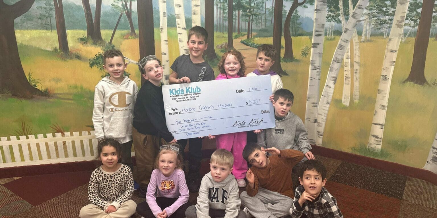 Children from EnrichEd Kids Klub smiling while holding a large donation check before a painted forest mural.