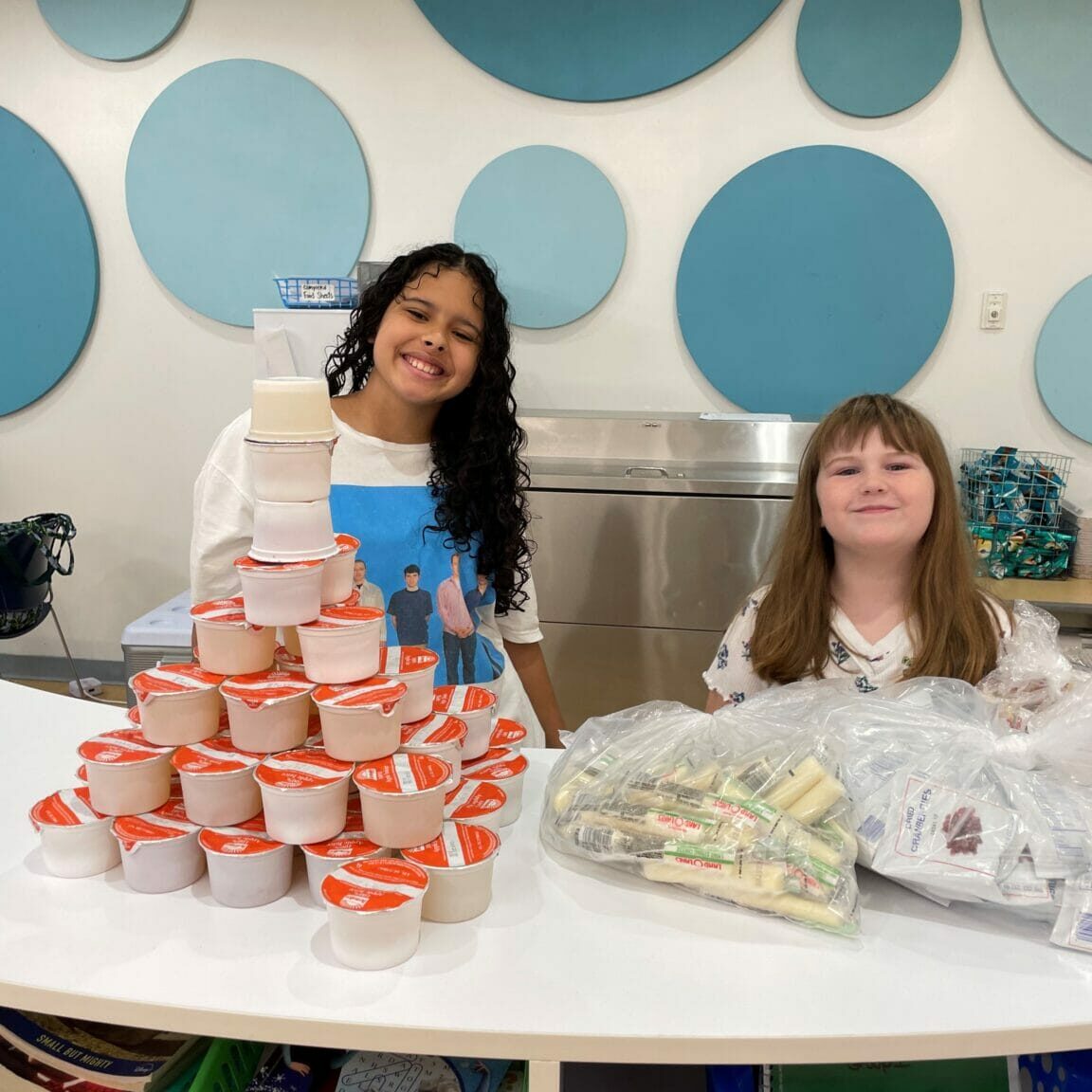 Two smiling girls at a snack station with stacked cups and cheese, blue circle wall decor — EnrichEd program space.