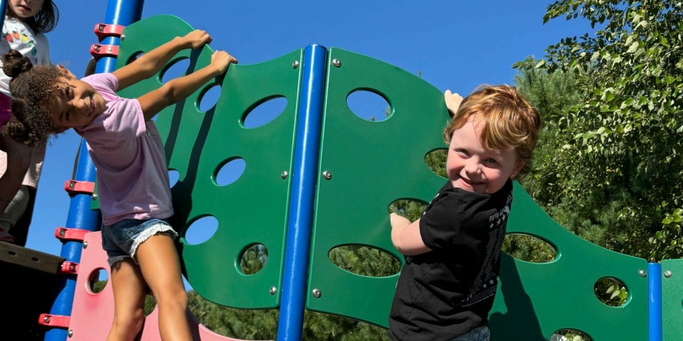Children climb a green playground panel under blue sky, smiling together, EnrichEd community enrichment and safe play