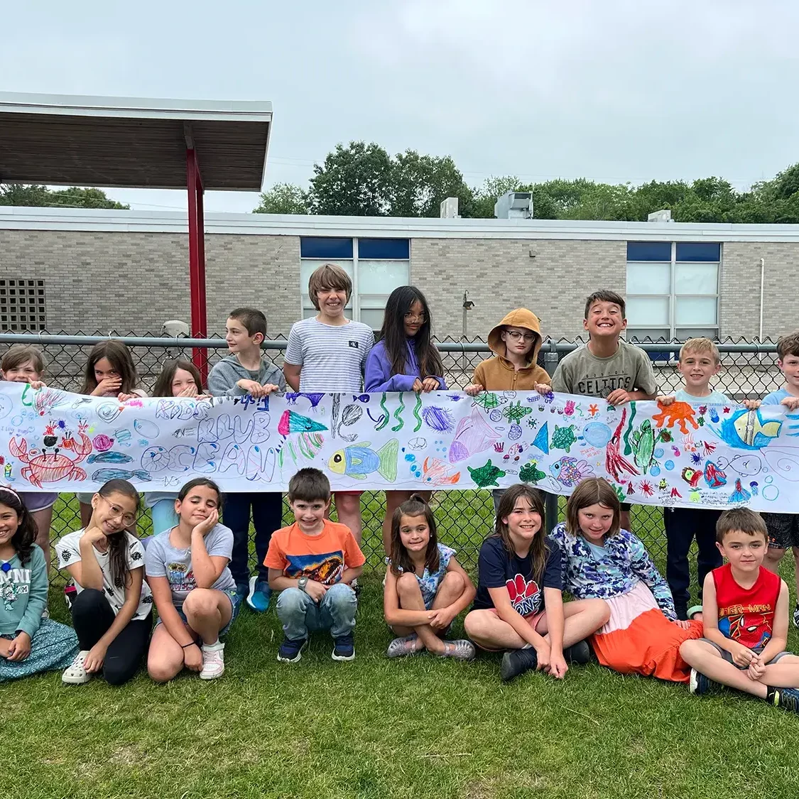 EnrichEd students smiling outdoors hold a long, hand-drawn ocean community art banner in front of a school fence.