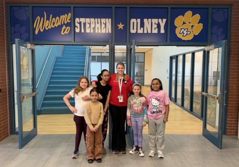Six students and a staff member smiling under 'Welcome to Stephen Olney' entrance, EnrichEd school community.