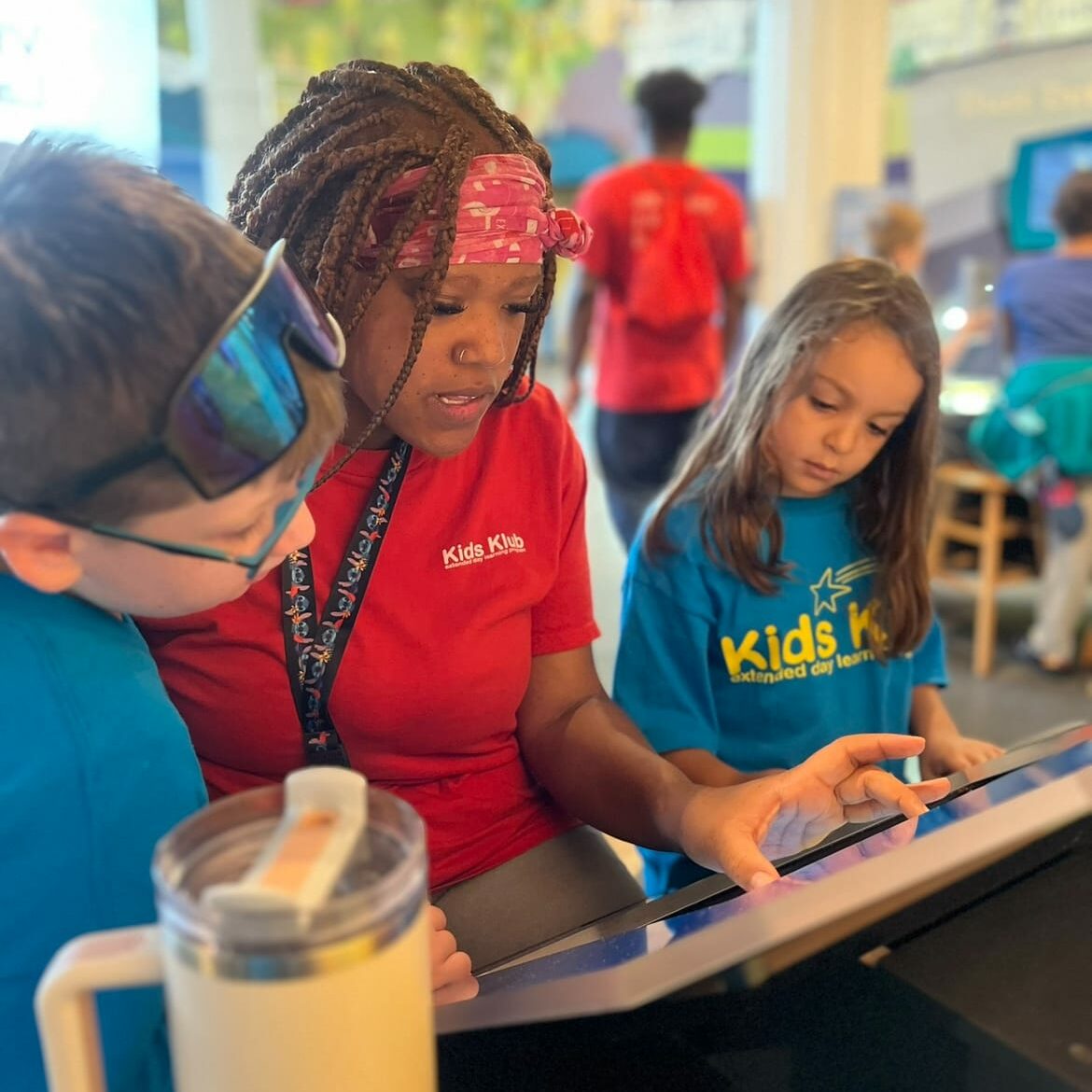 Leader in red Kids Klub shirt guiding two children on touchscreen tablet, EnrichEd enrichment in a supportive learning space