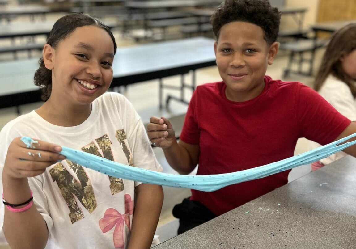 Two smiling students at a school cafeteria table stretching blue slime together during an EnrichEd activity